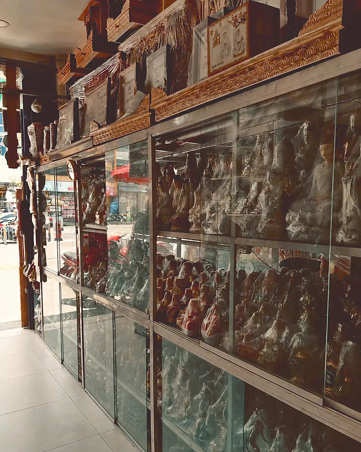 Bên trong cửa hàng đồ thờ của gia đình ở Chợ Lớn — tủ kính bày tượng, khung gỗ chạm trên nóc tủ